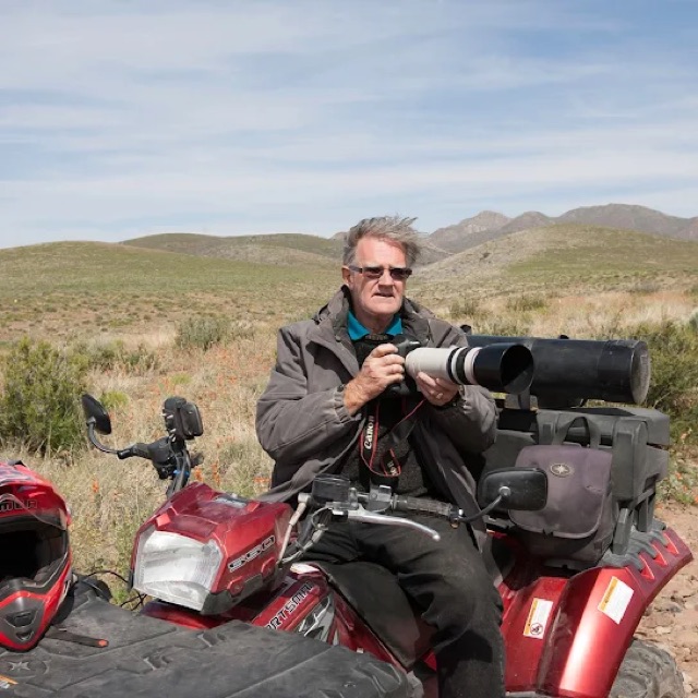 Sam sits on his red quad bike among desert hills, holding a Canon camera with a long white telephoto lens.
