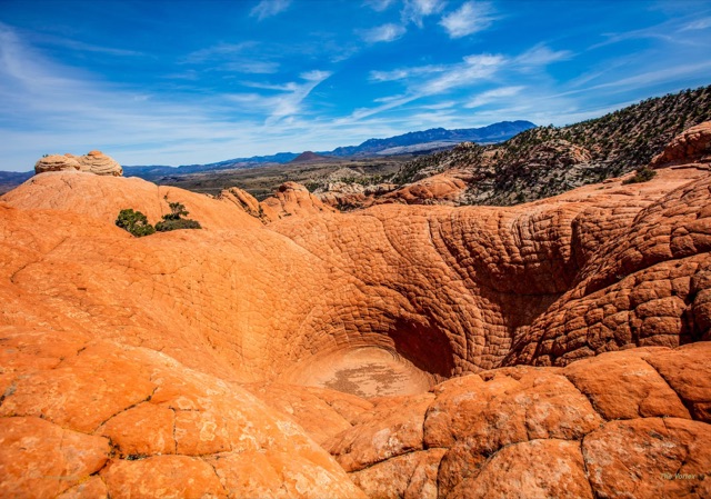 A deep bowl-shaped hollow swirled into orange slickrock, with mountains along the horizon.
